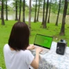 Person with shoulder-length hair typing on a silver laptop at a park table by a pond, surrounded by trees.